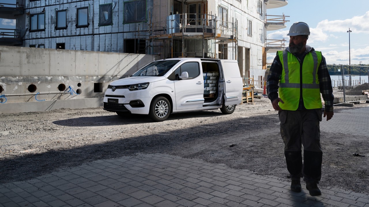 Ein Bauarbeiter mit weißem helm und neon-gelber Sicherheitsweste steht im Vordergrund. Hinter ihm eine Baustelle mit einem beladenen Proace City.