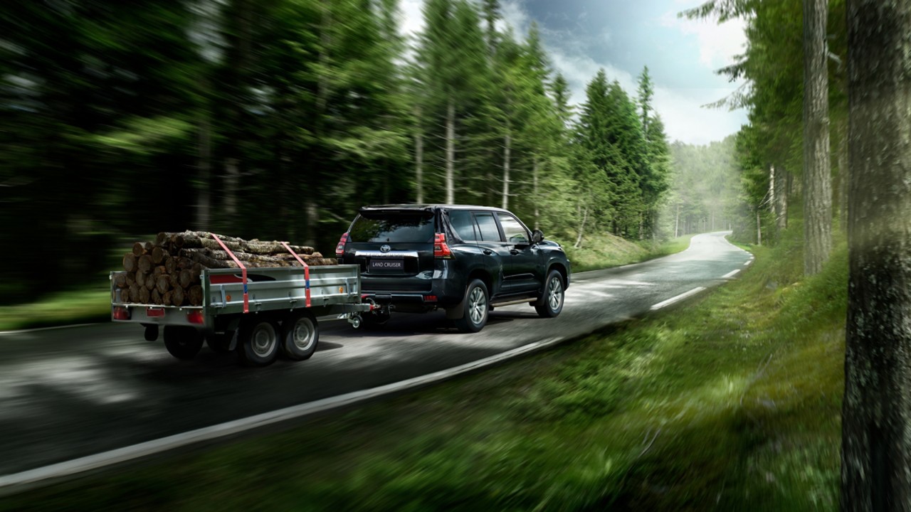 Ein schwarzer Landcruiser mit Anhänger, der mit Baumstämmen beladen ist auf einer Landstraße, die durch einen Wald führt.